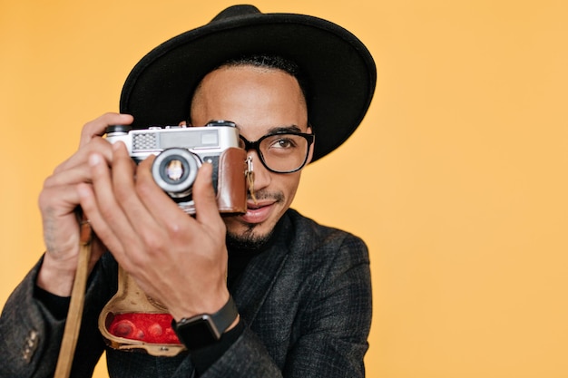 Dreamy male photographer taking pictures in studio with yellow interior. Indoor photo of joyful african guy with camera isolated on bright background.