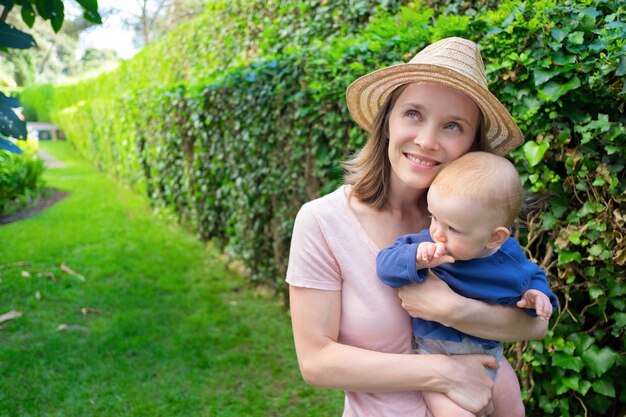 Dreamy mother in hat hugging cute infant, smiling and looking away. Adorable baby on mom hands with fist near cheek. Summer family time, garden 