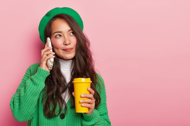 Мечтательная симпатичная азиатская девушка держит смартфон возле уха, наслаждается приятной беседой за чашкой кофе с собой, у нее длинные темные волосы, она одета в стильную зеленую одежду.