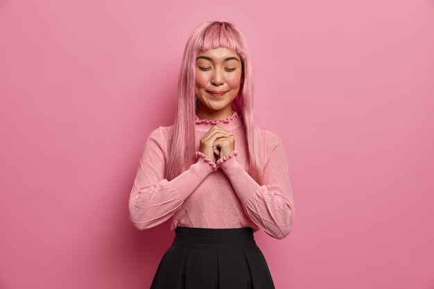 dreamy pleased Asian woman stands with eyes closed, smiles happily, has long pink hair, wears sweater