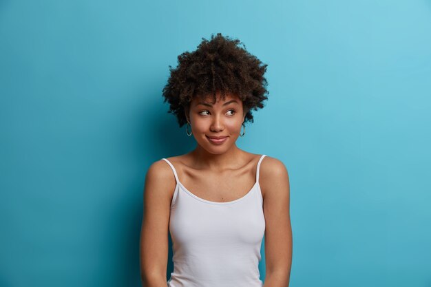 Dreamy positive dark skinned woman with Afro hair looks aside, has pleasant smile on face, dressed in casual vest, notices something pleasant, poses 