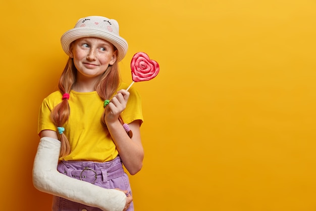 Dreamy positive freckled girl poses with big heart shaped lollipop, has sweet tooth, enjoys eating harmful food, holds delicious candy, wears fashionable summer outfit, has broken arm. Sugar addiction