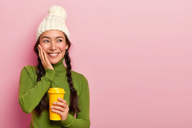 Dreamy positive teenage girl with plaits, touches cheeks, remembers something very pleasant during coffee break, holds takeaway cup of drink, wears winter hat and green poloneck, poses over rosy wall