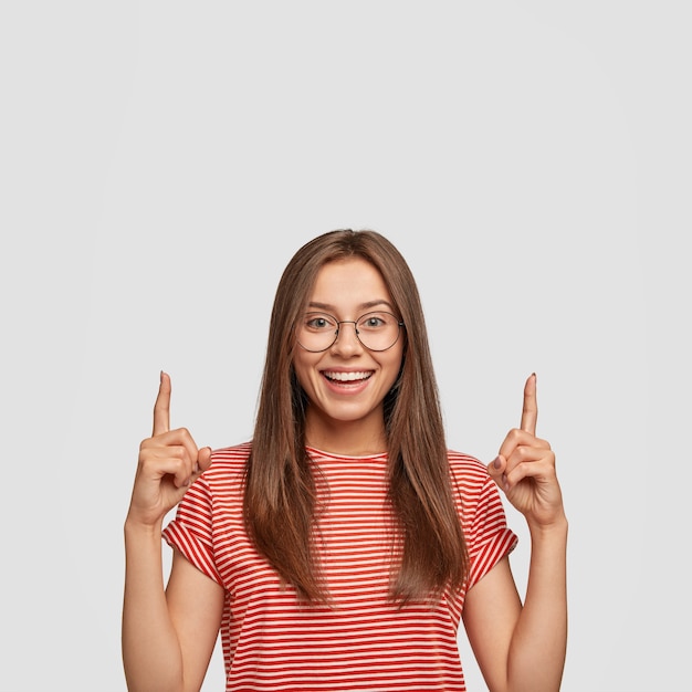 Free photo dreamy positive woman with satisfied expression, points with both index fingers upwards, thinks about new possibilities