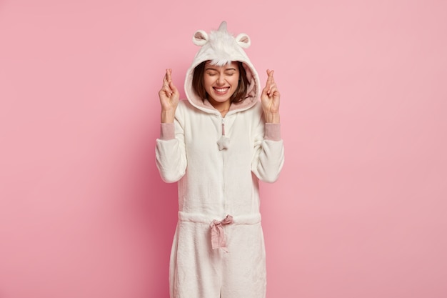 부드러운 kigurumi 의상을 입은 꿈꾸는 긍정적 인 젊은 여성, 손가락을 교차하고 행운을 믿습니다.