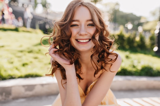Бесплатное фото Мечтательная рыжеволосая девушка позирует с искренней улыбкой на природе. великолепная рыжая кудрявая женщина, наслаждающаяся солнечным летним утром.