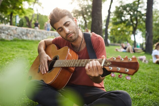 Мечтательный расслабленный мужчина играет на гитаре, сидит на траве в парке с инструментом