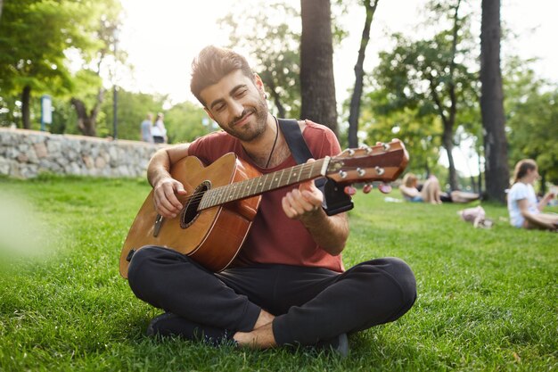 Мечтательный расслабленный мужчина играет на гитаре, сидит на траве в парке с инструментом