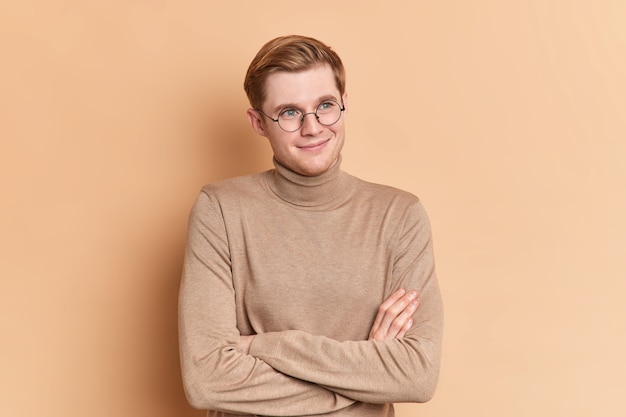 Dreamy satisfied blue eyed handsome male youngster stands crossed hands indoor looks into distance thinks about something pleasant wears casual beige turtleneck 