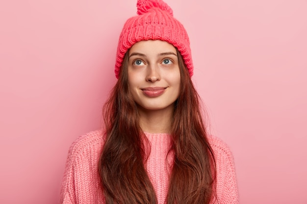 Dreamy satisfied brunette millennial girl looks above with pleasant thoughts, wears vivid knitted hat and sweater, models over pink background, has charming smile and tender look