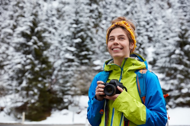 Dreamy satisfied European female traveler holds digital camera for making pictures, focused into distance