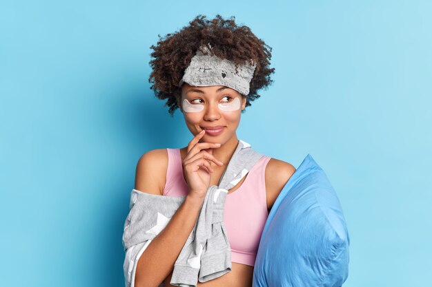 꿈꾸는 만족스러운 젊은 아프리카 계 미국인 여성은 눈 아래 콜라겐 뷰티 패치를 적용하고 좋은 분위기에서 깨어나 부드러운 베개를 보유하고 있습니다.