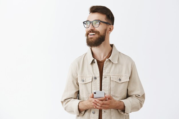 Dreamy smiling bearded man in glasses posing against the white wall