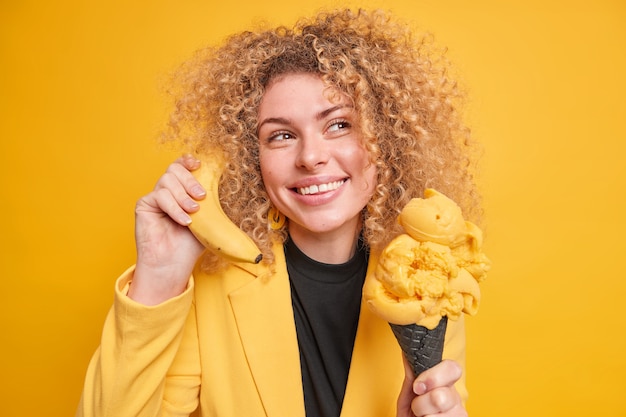 Dreamy smiling curly haired woman looks away thoughtfully holds cone ice cream eats frozen dessert holds banana near ear pretends having telephone talk being in good mood yellow  wall