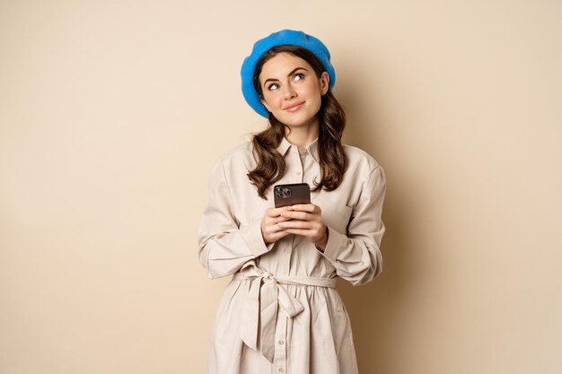 Dreamy smiling woman in stylish trenchcoat, looking up fantasizing while shopping on mobile phone app, using smartphone and thinking, standing over beige background
