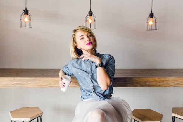 Dreamy stylish girl with blonde hair and pink lips sitting in a coffee shop with wooden chairs and table. She holds a cup of cofee