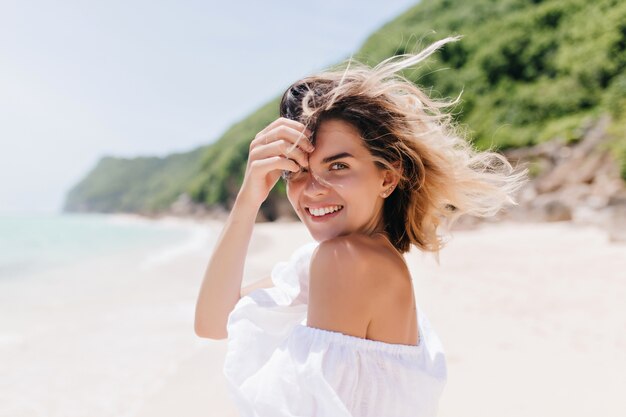 Dreamy tanned woman looking over shoulder while standing at sea coast. Adorable blonde woman posing with pleasure at tropical island in hot day.