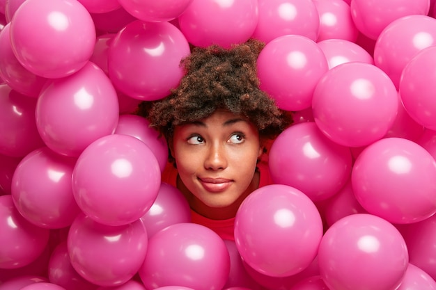 Dreamy thoughtful Afro American woman enjoys celebration, poses among pink balloons looks pensively aside makes plans for party.