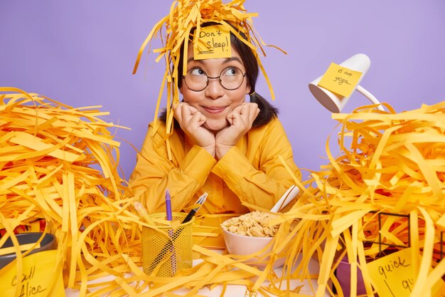 Dreamy thoughtful female office worker with paper wastes on head keeps hands under chin looks away pensively poses at desktop prepares project on purple backgound