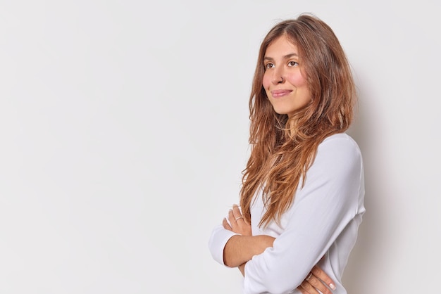 Dreamy thoughtful young woman stands with arms crossed smiles gently dressed in casual jumper concentrated into distance isolated over white background copy space for your promotional content