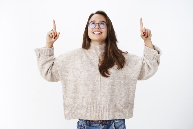 夢のようなスリルと幸せな若いブルネットの女性の学生は、灰色の壁に雪が降るのを見て、明るくのんきな笑顔で喜んで見上げて、頭と手を指して、眼鏡とセーターを上げています。