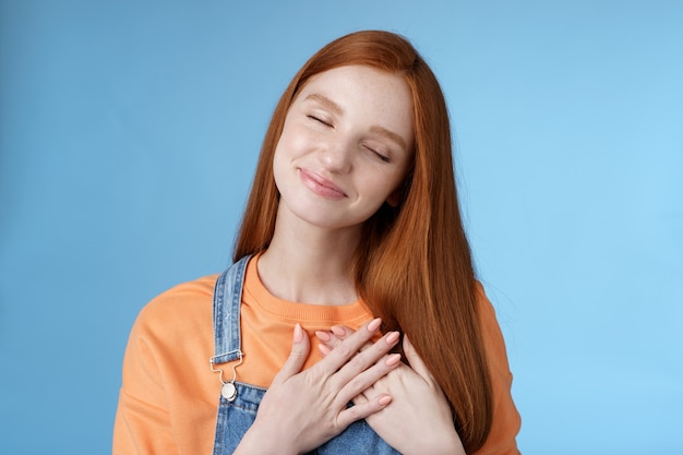 Dreamy touched romantic ginger girlfriend close eyes recalling heartwarming romance touch heart palms pressed chest smiling tenderly feel love care sympathy express affection, blue background