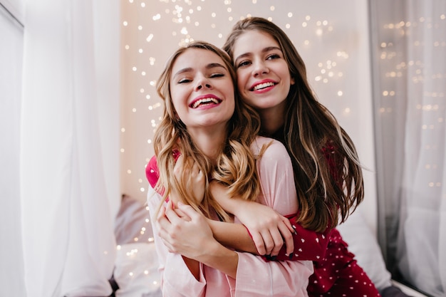 Dreamy white girl embracing sister and looking away with smile. Indoor photo of chilling female friends posing in pajamas.