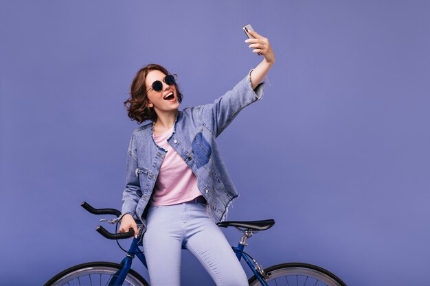 Мечтательная белая девушка с помощью телефона для селфи с новым велосипедом. Симпатичная кудрявая женщина в солнечных очках, стоящая возле велосипеда.
