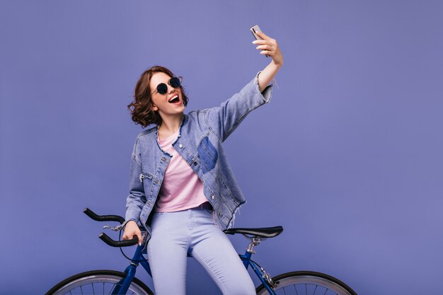 Dreamy white girl using phone for selfie with new bike. Winsome curly woman in sunglasses standing near bicycle.