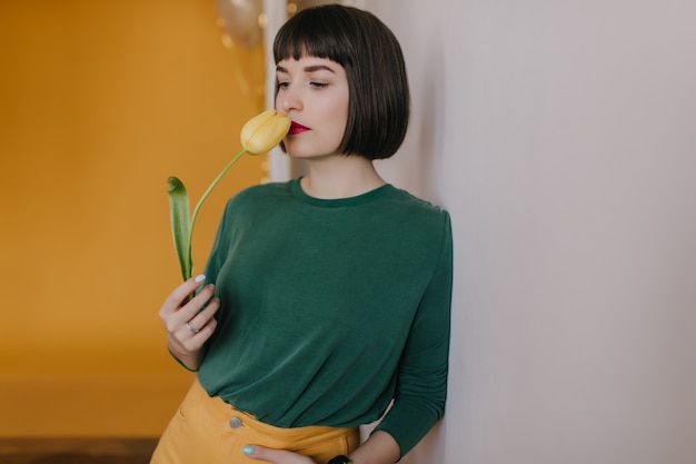 Dreamy woman in casual clothes sniffs yellow tulip. Indoor photo of adorable white girl with flower posing at home.