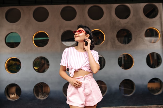 Мечтательная женщина, глядя вверх, позирует на фоне городских. Великолепная женщина в розовых шортах стоит на улице.