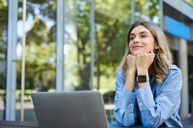 Free photo dreamy woman sitting with laptop outdoors on street smiling businesswoman daydreaming imaging smth w