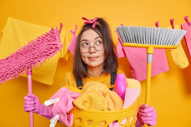 Dreamy woman stands with cleaning tools concentrates thoughtfully above thinks what to do after finishing work about house poses near basin of laundry haging clothesline behind. Domestic chores