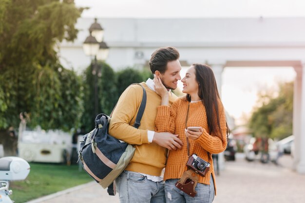 Dreamy woman in stylish orange sweater holding smartphone and gently touching boyfriend's face