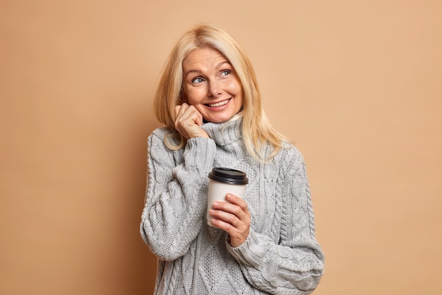 Dreamy wrinkled woman pensioner with blonde hair minimal makeup dressed in warm grey sweater dreams about something pleasant and drinks coffee.