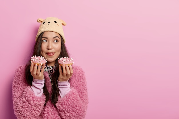 Dreamy young Asian lady holds two delicious sweet donuts, licks lips, wants to eat high calories food, breaks diet, dressed in warm clothes and headgear