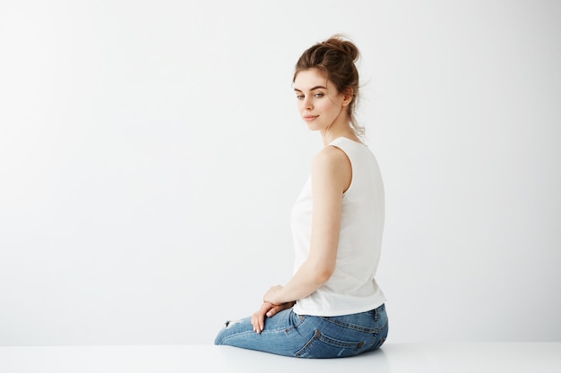 Dreamy young beautiful brunette woman with bun smiling thinking sitting over white background.