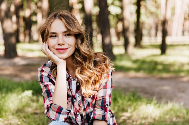 Dreamy young lady in casual shirt sitting in park and looking. Romantic woman with wavy hair chilling in warm summer day.