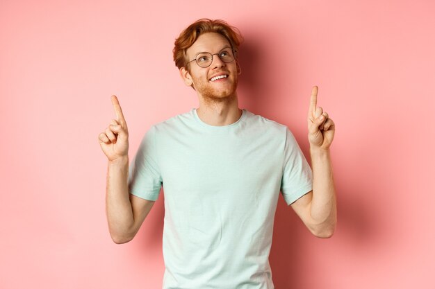 Dreamy young man with red hair and beard pointing fingers and looking cheerful up admire something s...