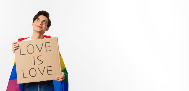 Free photo dreamy young queer person smiling and looking at upper left corner holding love is love sign for pride parade wearing rainbow flag white background