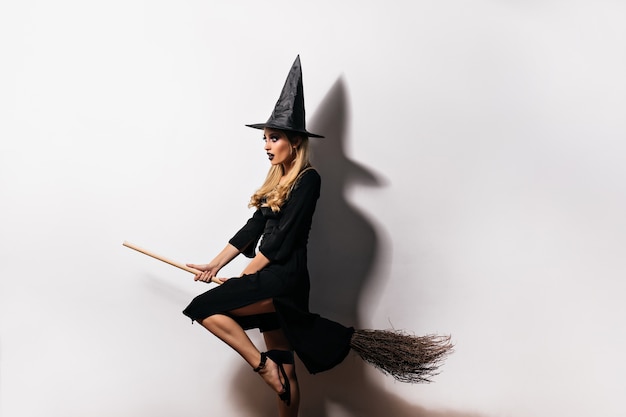 Dreamy young witch flying on broom in halloween. Indoor shot of debonair blonde wizard posing on white wall.