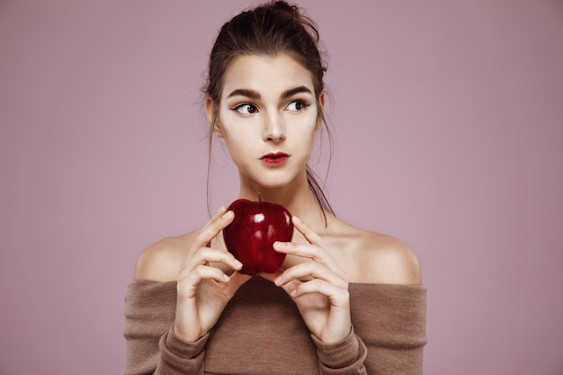 Free photo dreamy young woman holding red apple looking in side on pink