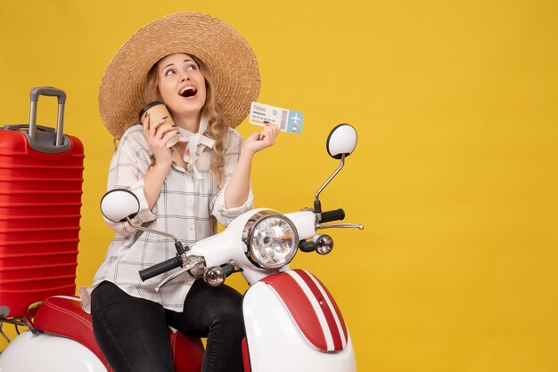 Dreamy young woman wearing hat and sitting on motorcycle and holding coffee and ticket on yellow 