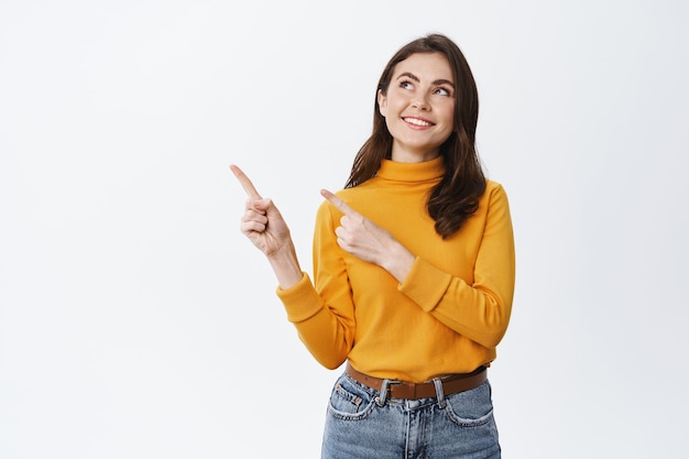 Dreamy young woman with natural beauty face, looking and pointing at upper left corner, imaging something, gazing at something interesting, standing on white wall