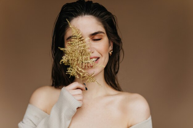 Dreamy young woman with nude makeup posing with plant. Close-up portrait of ecstatic black-haired girl chilling .