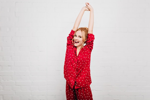 Dreamy young woman with pleased face expression stretching in morning. Cute european girl enjoying photoshoot in red night-suit.