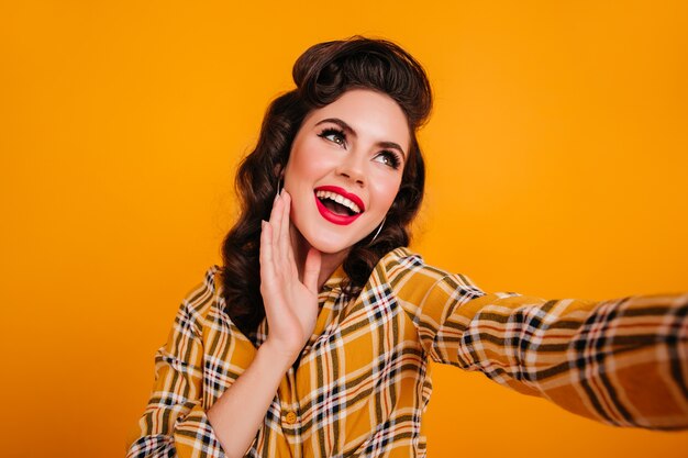 Dreamy young woman with red lips taking selfie. Studio shot of pinup lady in checkered shirt posing on yellow background.