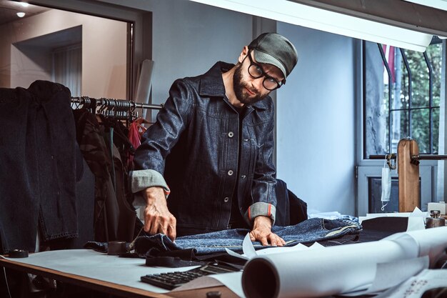 Dressmaker is measuring fabric  with meter  in his studio. Man is looking to the camera. He is wearing denim, cap and glasses. There are a lot of sewing tools at the background.