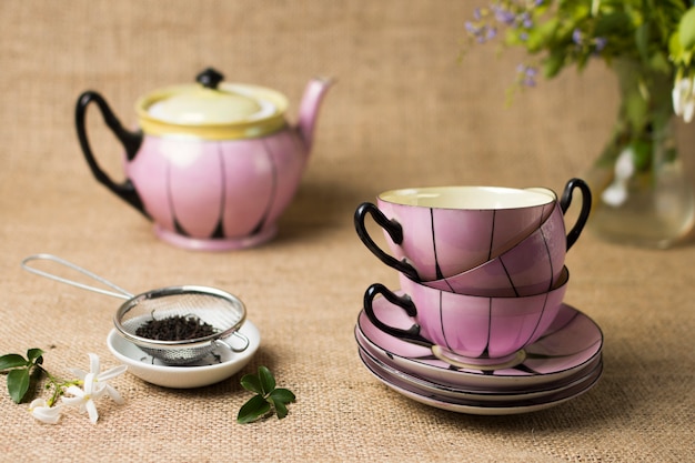 Сушеный черный чай с цветами и стопкой керамической чашки с блюдцами на джутовой скатерти