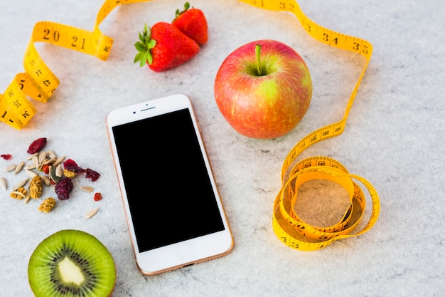Бесплатное фото Сухофрукты; яблоко; наполовину киви; клубника; измерительная лента и смартфон на сером текстурированном фоне